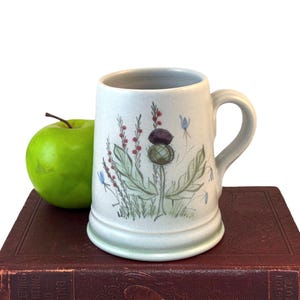 May include: A white ceramic mug with a blue and green thistle design. The mug has a handle and is sitting on a brown book.