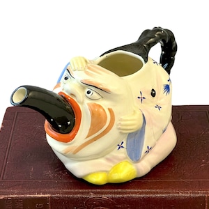May include: A ceramic teapot shaped like a person's head with a black spout and a blue and white patterned design. The teapot is sitting on a brown book.