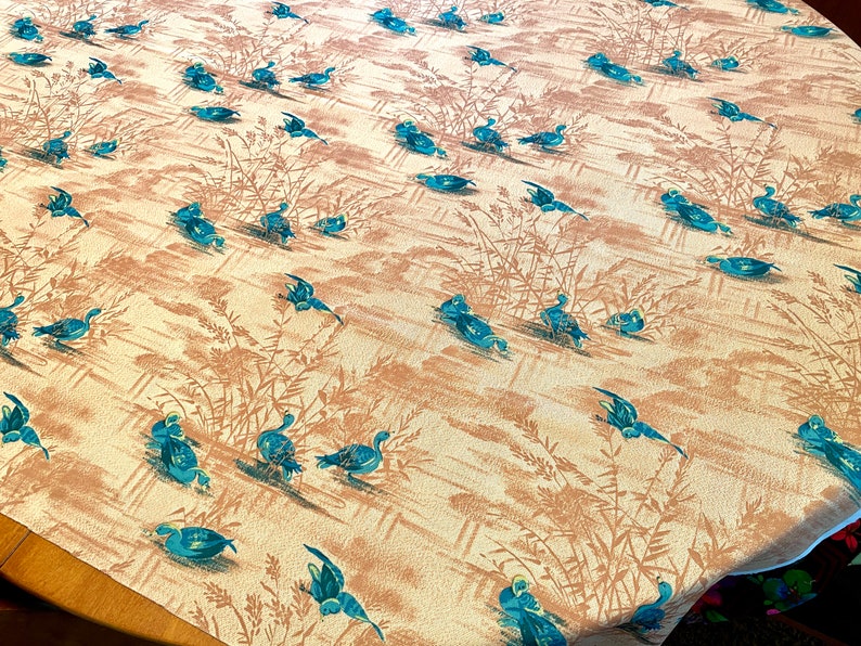 May include: A vintage tablecloth with a repeating pattern of blue ducks swimming in a pond with brown reeds. The background is a light beige color.