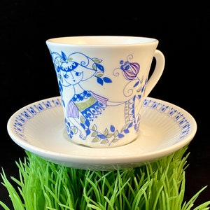 Może przedstawiać: Biały ceramiczny kubek i spodek z niebieskim i zielonym wzorem kwiatowym przedstawiającym stylizowaną kobietę. Kubek i spodek mają niebieską i białą obwódkę.