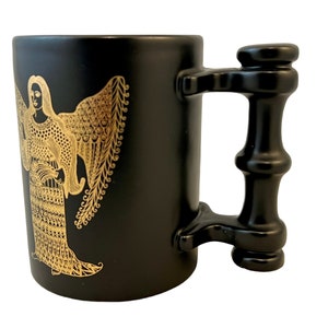 K&ouml;nnte beinhalten: Schwarze Keramiktasse mit einem goldenen Engelmotiv. Die Tasse hat einen einzigartigen schwarzen Griff mit zylindrischer Form.