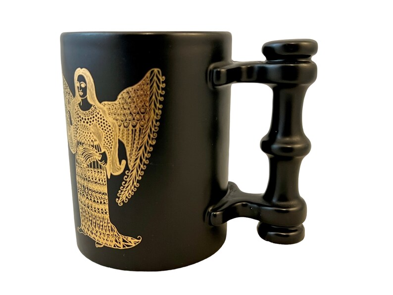 K&ouml;nnte beinhalten: Schwarze Keramiktasse mit einem goldenen Engelmotiv. Die Tasse hat einen einzigartigen schwarzen Griff mit zylindrischer Form.
