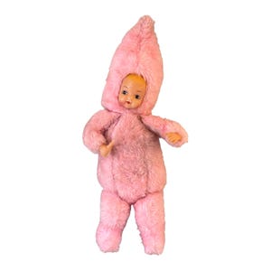 Op de afbeelding: Een vintage pop met een roze, fuzzy jumpsuit en bijpassende capuchon. De pop heeft een geschilderd gezicht met details en staat rechtop. De outfit bedekt de armen en benen van de pop.