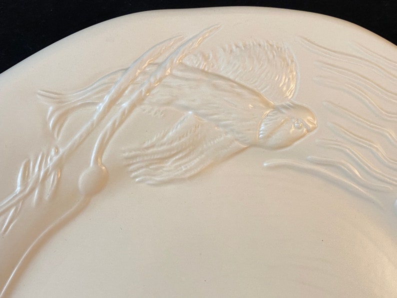 K&ouml;nnte beinhalten: Wei&szlig;e Keramik-Servierplatte mit einem erhabenen Design eines Fisches, der durch Schilf schwimmt. Der Fisch ist mit Flossen und Schuppen detailliert.