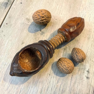 May include: A dark brown wooden nutcracker with a screw-style mechanism and a rounded bowl-shaped end, accompanied by three whole walnuts with textured brown shells. The nutcracker features a polished finish and a threaded central handle.