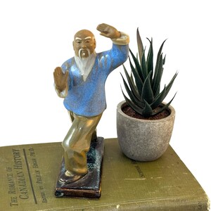 May include: A ceramic figurine of an elderly man in a martial arts pose, wearing a blue top and tan pants. The figurine is on a rectangular base. A small potted succulent sits next to the figurine on a vintage book.