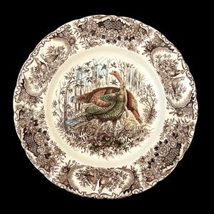 May include: A round ceramic dinner plate with a cream-colored base featuring a central illustration of a turkey in a forest setting. The scalloped rim is decorated with a brown transferware pattern of floral motifs and small game birds.