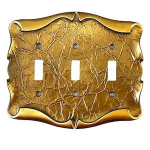Puede incluir: Una placa de interruptor de luz decorativa de color dorado con una superficie texturizada arrugada y tres interruptores de palanca. La placa tiene un marco decorativo ornamentado.