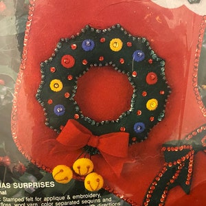 May include: A red felt stocking with a green felt wreath decorated with colorful sequins and buttons. The stocking also has a red felt bow and three yellow felt bells.