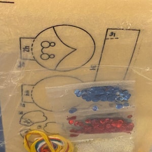 May include: A craft kit with a pattern for making a felt ornament. The kit includes blue and red sequins, yellow embroidery floss, and a white felt sheet.