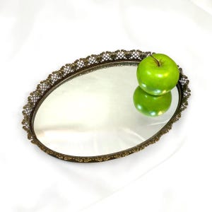 Puede incluir: Una bandeja ovalada con una base de espejo reflectante y un borde decorativo de metal en tono bronce que presenta un intrincado patrón de celosía y volutas. Una manzana de color verde brillante descansa sobre la superficie del espejo.