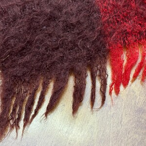 May include: Close-up of a textured, fringed scarf. The scarf features sections of deep burgundy and vibrant red, with a hint of pink. The fringed edges add a decorative element to the cozy, woven textile.