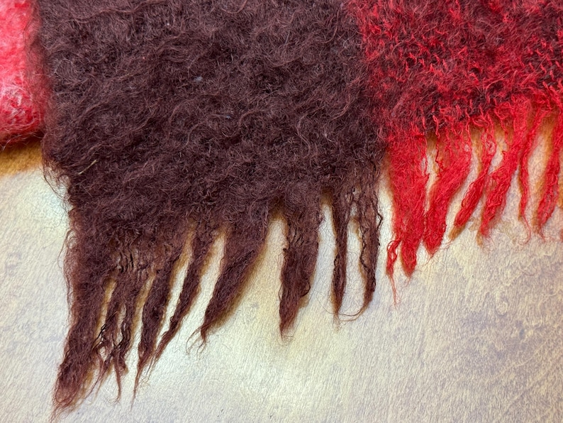 May include: Close-up of a textured, fringed scarf. The scarf features sections of deep burgundy and vibrant red, with a hint of pink. The fringed edges add a decorative element to the cozy, woven textile.