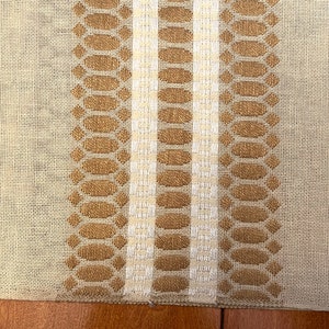 May include: A beige linen table runner with two brown and white woven stripes. The stripes feature a repeating pattern of oval shapes.