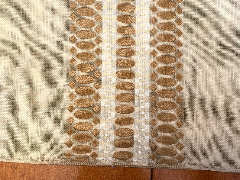 May include: A beige linen table runner with two brown and white woven stripes. The stripes feature a repeating pattern of oval shapes.