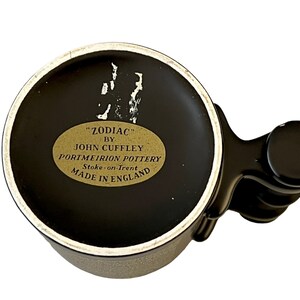 K&ouml;nnte beinhalten: Schwarze und wei&szlig;e Keramiktasse mit schwarzem Henkel. Die Tasse hat ein goldenes Etikett mit der Aufschrift "ZODIAC" BY JOHN CUFFLEY PORTMEIRION POTTERY Stoke-on-Trent MADE IN ENGLAND.