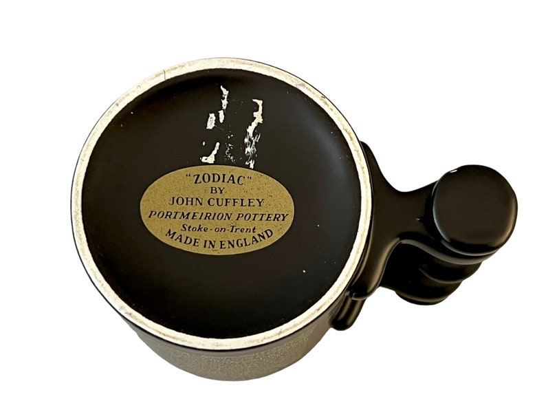 K&ouml;nnte beinhalten: Schwarze und wei&szlig;e Keramiktasse mit schwarzem Henkel. Die Tasse hat ein goldenes Etikett mit der Aufschrift "ZODIAC" BY JOHN CUFFLEY PORTMEIRION POTTERY Stoke-on-Trent MADE IN ENGLAND.
