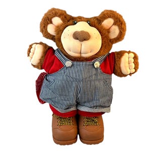 以下が含まれることがあります： 赤いシャツと白のストライプの青いオーバーオールを着た茶色のぬいぐるみクマ。クマは茶色の毛皮と茶色の鼻をしています。クマは茶色のブーツを履いています。