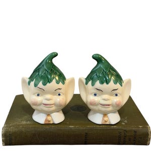 May include: Two ceramic elf figurines featuring cream-colored faces with blue eyes, pink cheeks, and pointed ears. Each elf wears a green leaf-patterned hat and a white collar with a small tan accent. The figures have a glossy finish and expressive faces.