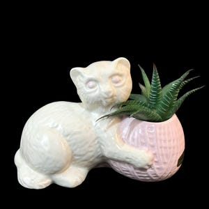 Puede incluir: Maceta de cerámica blanca con forma de gato con una maceta de bola de lana rosa en su interior. La maceta de bola de lana tiene una planta suculenta verde que crece en su interior.
