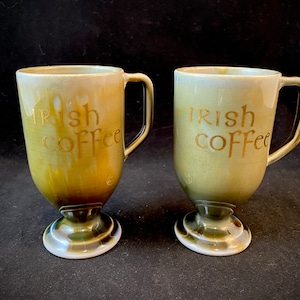 Puede incluir: Dos tazas de cerámica verde y blanca con letras doradas que dicen "Irish Coffee". Las tazas tienen una base de pedestal.
