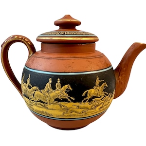 Puede incluir: Tetera de color terracota con una banda negra que representa una escena de caza dorada. La tapa tiene un diseño geométrico dorado y negro. El asa y el pico también son de color terracota con detalles dorados.