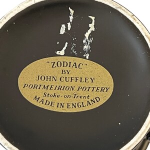 K&ouml;nnte beinhalten: Nahaufnahme des Bodens einer schwarzen Keramiktasse mit einem goldenen Etikett mit der Aufschrift "ZODIAC" von John Cuffley, Portmeirion Pottery, Stoke-on-Trent, Hergestellt in England.