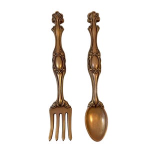 Puede incluir: Un par de utensilios de cocina decorativos para pared que consisten en un tenedor y una cuchara. Ambos artículos presentan un acabado en bronce metálico con detalles ornamentados en relieve en los mangos y un motivo en forma de abanico.