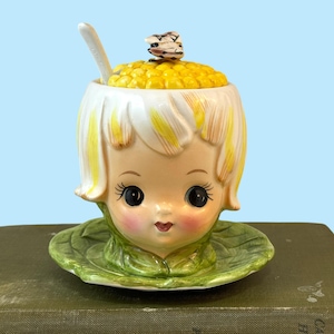 以下が含まれることがあります： 黄色い髪の女の子の頭の形をしたヴィンテージのセラミック製の砂糖入れ。コーンの蓋が付いています。緑の葉の形をした受け皿の上に置かれています。