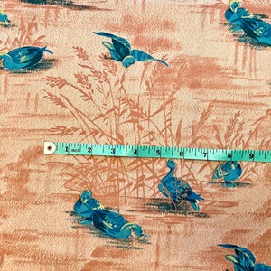 May include: A beige fabric with a repeating pattern of blue ducks and brown reeds. A measuring tape is placed across the fabric, showing a measurement of 11 inches.