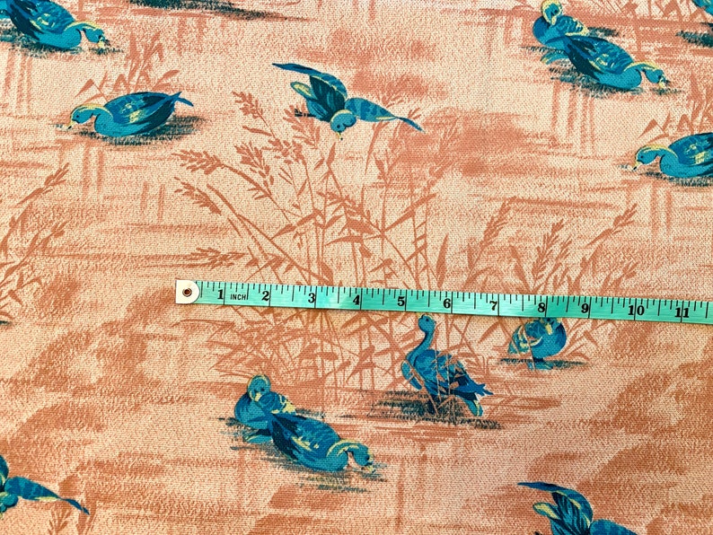 May include: A beige fabric with a repeating pattern of blue ducks and brown reeds. A measuring tape is placed across the fabric, showing a measurement of 11 inches.