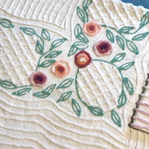 May include: A white quilted throw blanket with a floral design embroidered in green and pink. The blanket has a textured surface and a scalloped edge.