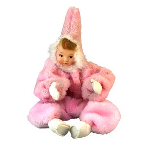 May include: A vintage doll with a baby face, wearing a pink fuzzy hooded outfit with white trim. The doll has white mittens and white booties. The doll is sitting down.