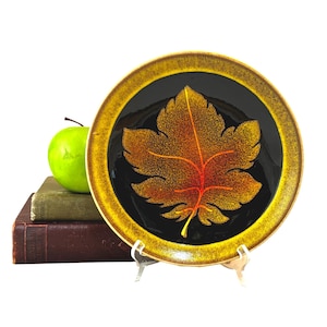 以下が含まれることがあります： 黒い背景に金色の葉のデザインが施された、装飾的なセラミックプレート。葉は赤で縁取られており、斑点のある質感があります。