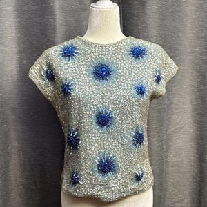 May include: A silver and blue beaded top with short sleeves and a round neckline. The top is covered in small silver beads and features a pattern of blue flowers.