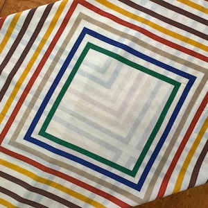 May include: A white fabric with a geometric pattern of brown, beige, red, and yellow stripes. The stripes are arranged in a diagonal pattern, creating a diamond shape in the center. The diamond shape is outlined with a blue and green border.