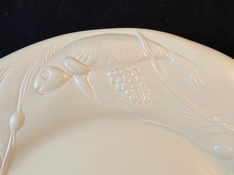 K&ouml;nnte beinhalten: Wei&szlig;e Keramik-Servierplatte mit einem gepr&auml;gten Design eines Fisches und Weizen&auml;hren.