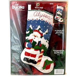 Peut inclure: Un kit de chaussette en feutre Bucilla représentant le Père Noël en train de livrer du courrier. La chaussette est bleue et blanche avec une canne en sucre rouge et blanche. Le kit comprend des instructions et des matériaux pour créer une chaussette personnalisée. La chaussette est étiquetée "Santa's Mail" et "Alyssa".