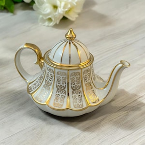 Op de afbeelding: Witte theepot met een gouden deksel en ingewikkelde gouden ontwerpen. De theepot heeft een rond lichaam en een tuit aan de zijkant.