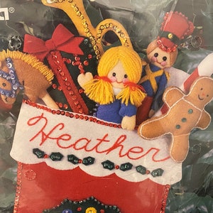 May include: A red felt stocking with a white cuff and the name "Heather" embroidered on it. The stocking is filled with felt toys, including a gingerbread man, a toy soldier, a rocking horse, and a doll.