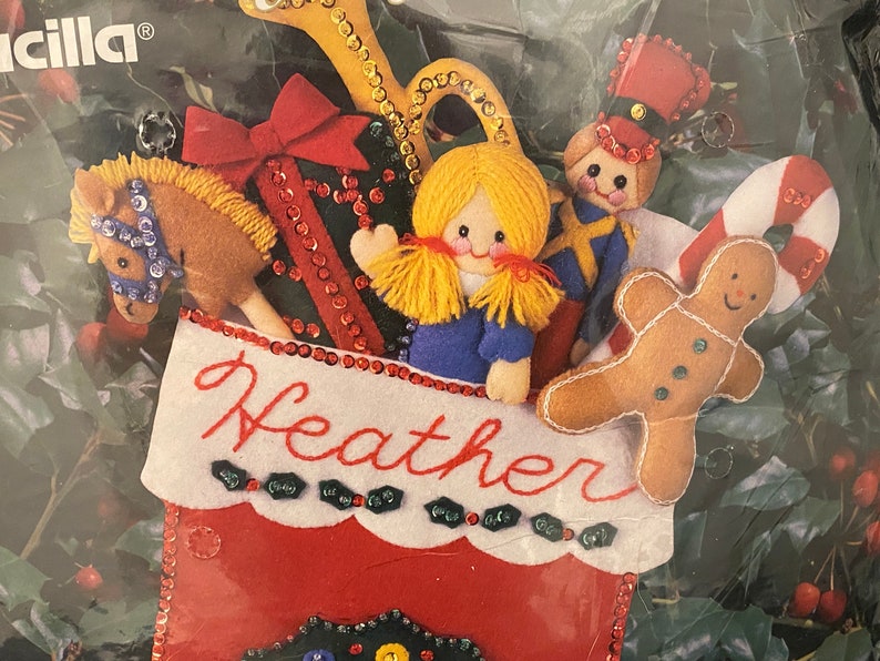 May include: A red felt stocking with a white cuff and the name "Heather" embroidered on it. The stocking is filled with felt toys, including a gingerbread man, a toy soldier, a rocking horse, and a doll.