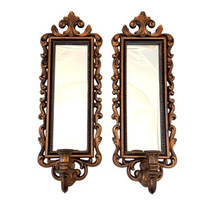 May include: A pair of rectangular wall mirrors with ornate bronze-colored frames featuring scrollwork and fleur-de-lis accents at the top. Each mirror includes a built-in candle holder at the base, extending outward from the bottom of the frame.