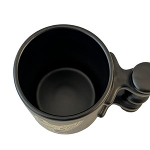 K&ouml;nnte beinhalten: Schwarze Keramiktasse mit einem einzigartigen Griffdesign. Der Griff hat einen abgerundeten Knopf am Ende und eine strukturierte Oberfl&auml;che. Die Tasse ist leer.