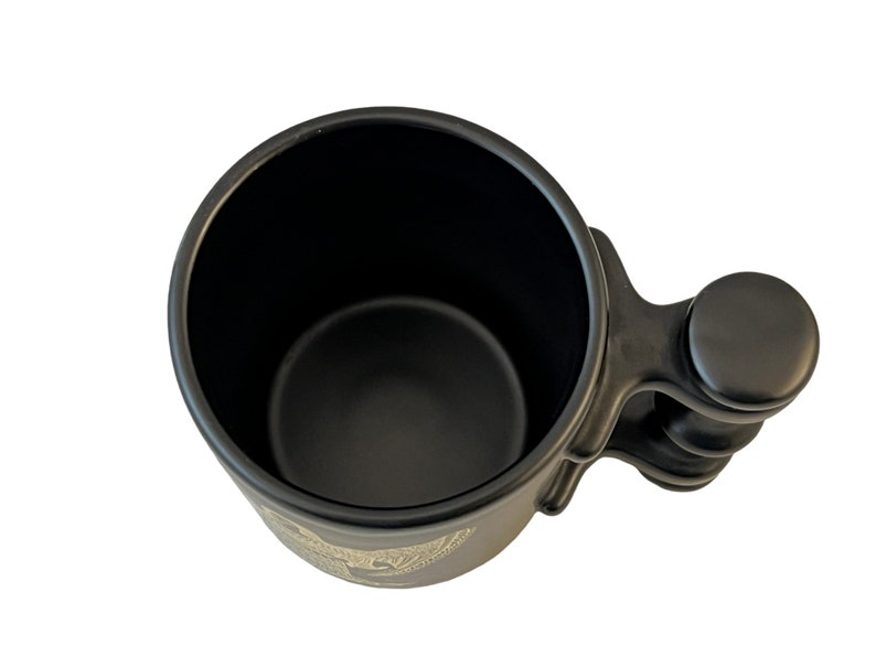 K&ouml;nnte beinhalten: Schwarze Keramiktasse mit einem einzigartigen Griffdesign. Der Griff hat einen abgerundeten Knopf am Ende und eine strukturierte Oberfl&auml;che. Die Tasse ist leer.
