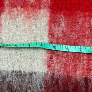 May include: Close-up of a red, white, and gray plaid fabric with a green measuring tape laid across it. The tape shows measurements in inches, with the fabric's texture visible. The fabric appears to be a blanket or throw.