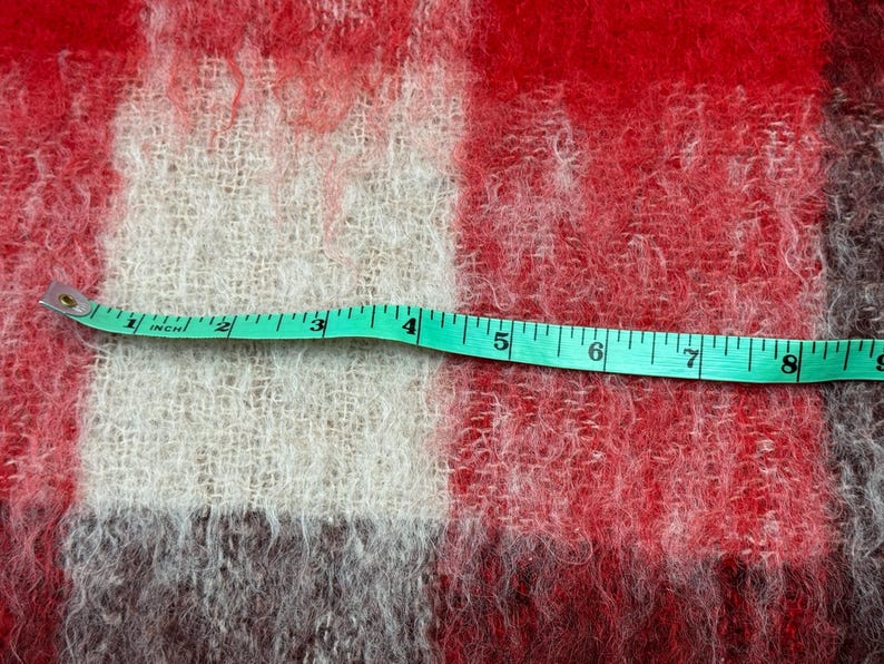 May include: Close-up of a red, white, and gray plaid fabric with a green measuring tape laid across it. The tape shows measurements in inches, with the fabric's texture visible. The fabric appears to be a blanket or throw.