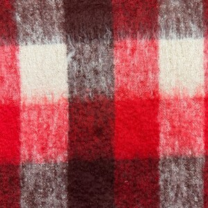 May include: Close-up of a woven fabric with a plaid pattern. The design features large squares in shades of red, brown, gray, and off-white. The fabric has a fuzzy, textured appearance, suggesting a warm, cozy material.