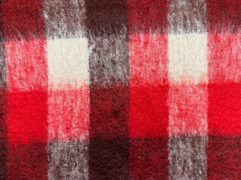 May include: Close-up of a woven fabric with a plaid pattern. The design features large squares in shades of red, brown, gray, and off-white. The fabric has a fuzzy, textured appearance, suggesting a warm, cozy material.