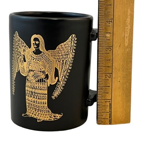 K&ouml;nnte beinhalten: Schwarze Keramiktasse mit einem goldenen Engelmotiv. Der Engel ist mit Fl&uuml;geln und einem langen, flie&szlig;enden Gewand dargestellt. Die Tasse hat einen Henkel.