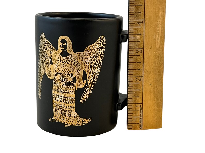 K&ouml;nnte beinhalten: Schwarze Keramiktasse mit einem goldenen Engelmotiv. Der Engel ist mit Fl&uuml;geln und einem langen, flie&szlig;enden Gewand dargestellt. Die Tasse hat einen Henkel.